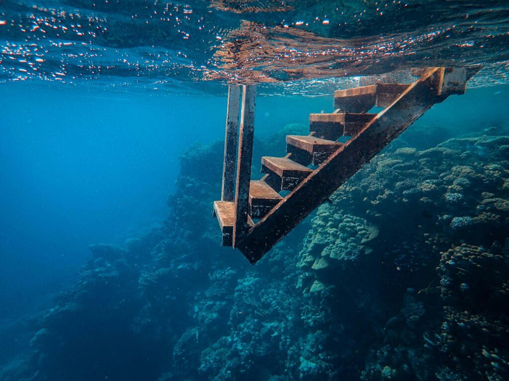 Underwater view of metal stairs leading to a vibrant coral reef in clear blue water.