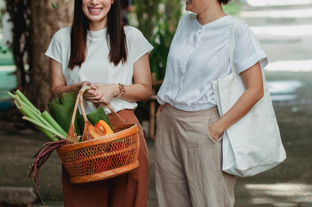 Crop unrecognizable glad woman with hand in pocket near partner with basket full of vegetables conversing in town