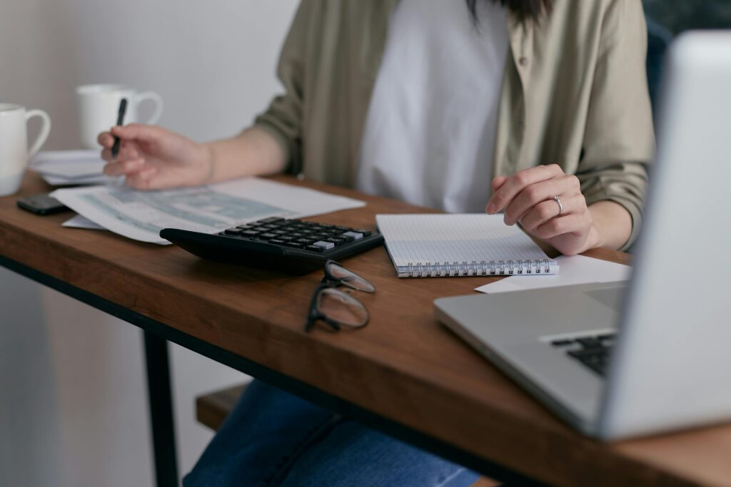 A woman manages finances at home, using a laptop and calculator on a wooden desk.