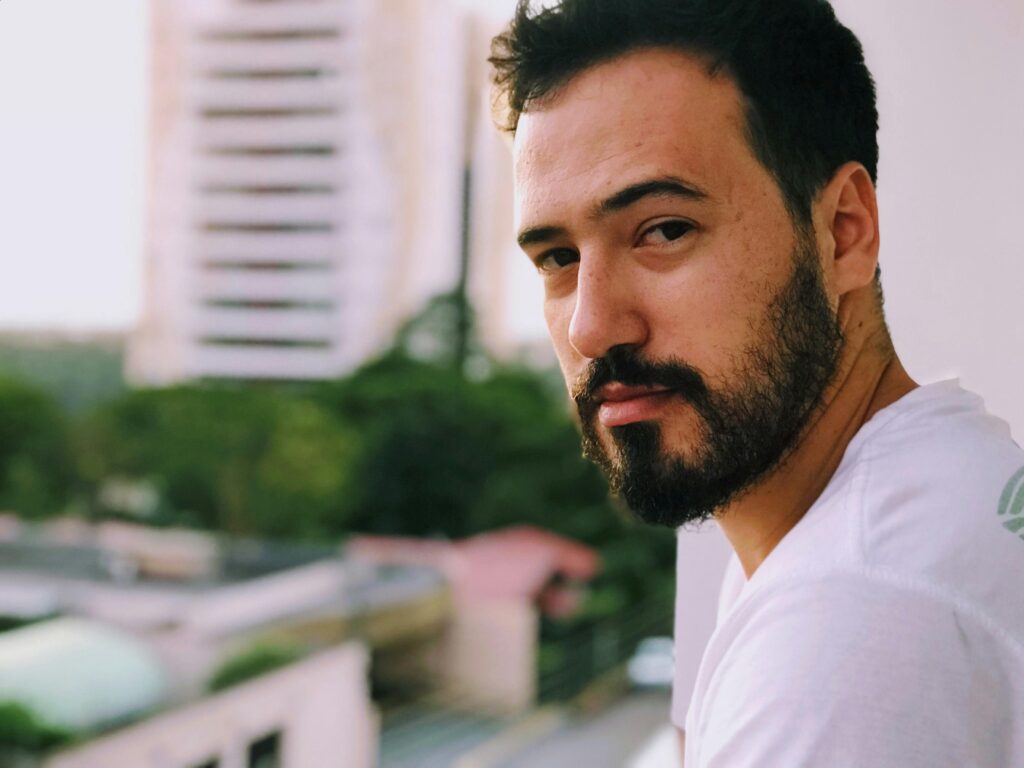 A serious-looking man with a beard stands on a balcony with blurred city buildings in the background.